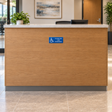 Blue 6 x 3 inch Please Ask for Assistance engraved sign with ISA wheelchair symbol mounted on the front of a reception desk; SKU AFA-0603-BLU.